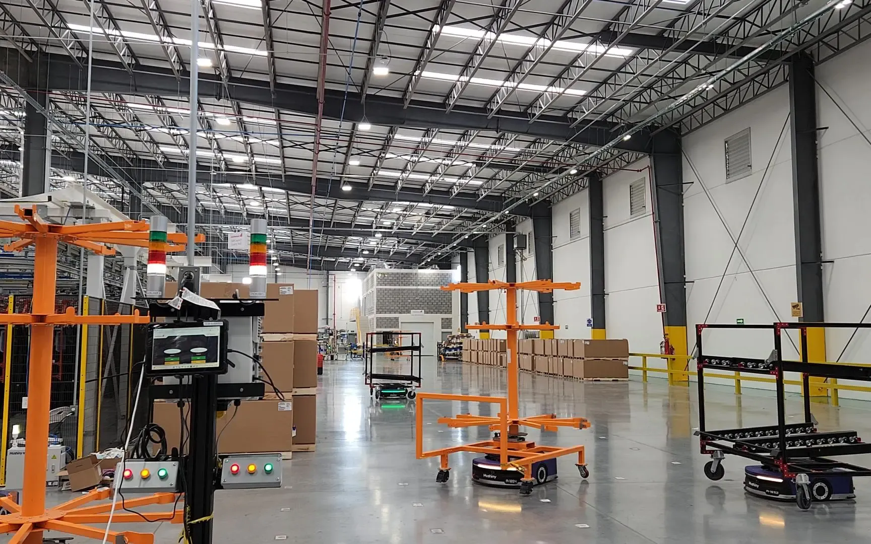 Control pedestal with a touchscreen HMI, pushbutton stations, and stack lights overseeing autonomous mobile robots that carry orange tooling racks across a warehouse floor
