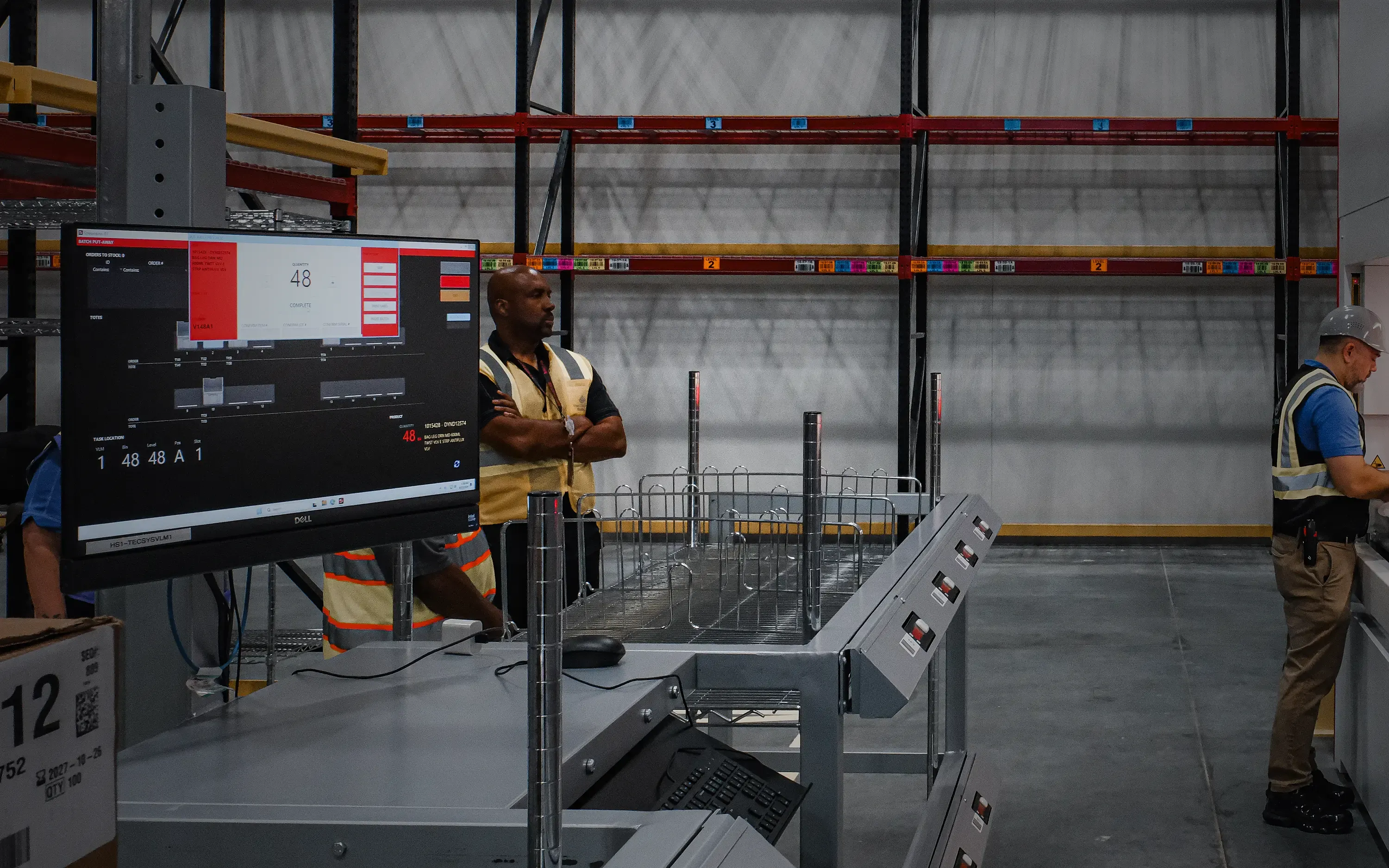 SRT batch put-away screen on a workstation monitor showing task location, quantity, and tote assignments while an operator picks at the vertical lift module access window behind it