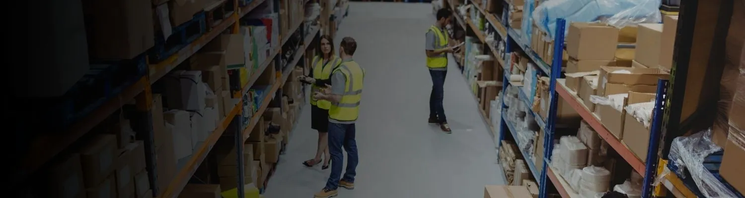 Warehouse staff in high-visibility vests working in an aisle lined with shelves of cartons and cases
