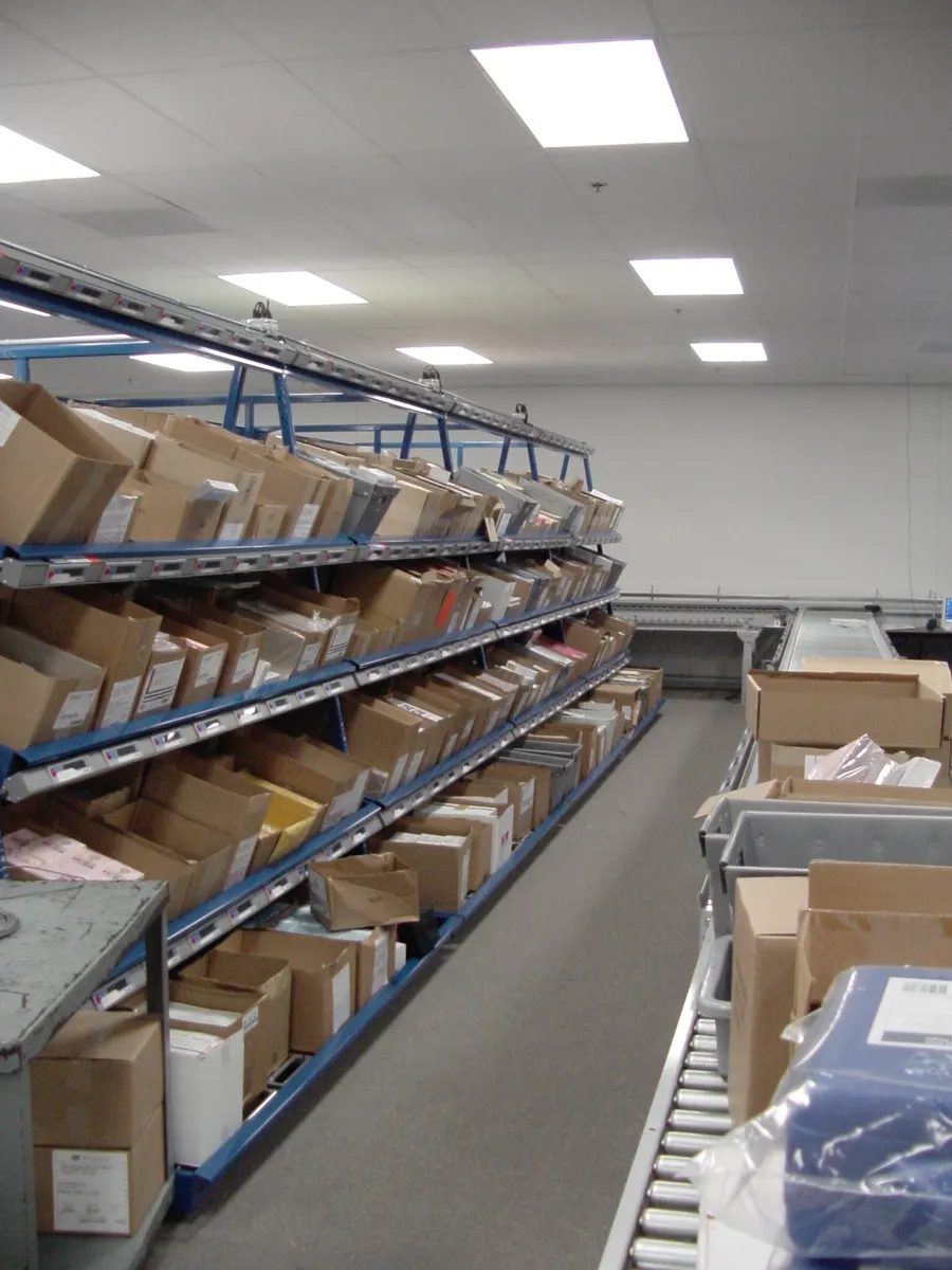 Carton flow rack aisle lined with pick-to-light displays above each pick face, with a roller conveyor running alongside