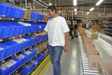 Operator walking a pick-to-light bin aisle and reaching for an item at an illuminated location beside a takeaway conveyor