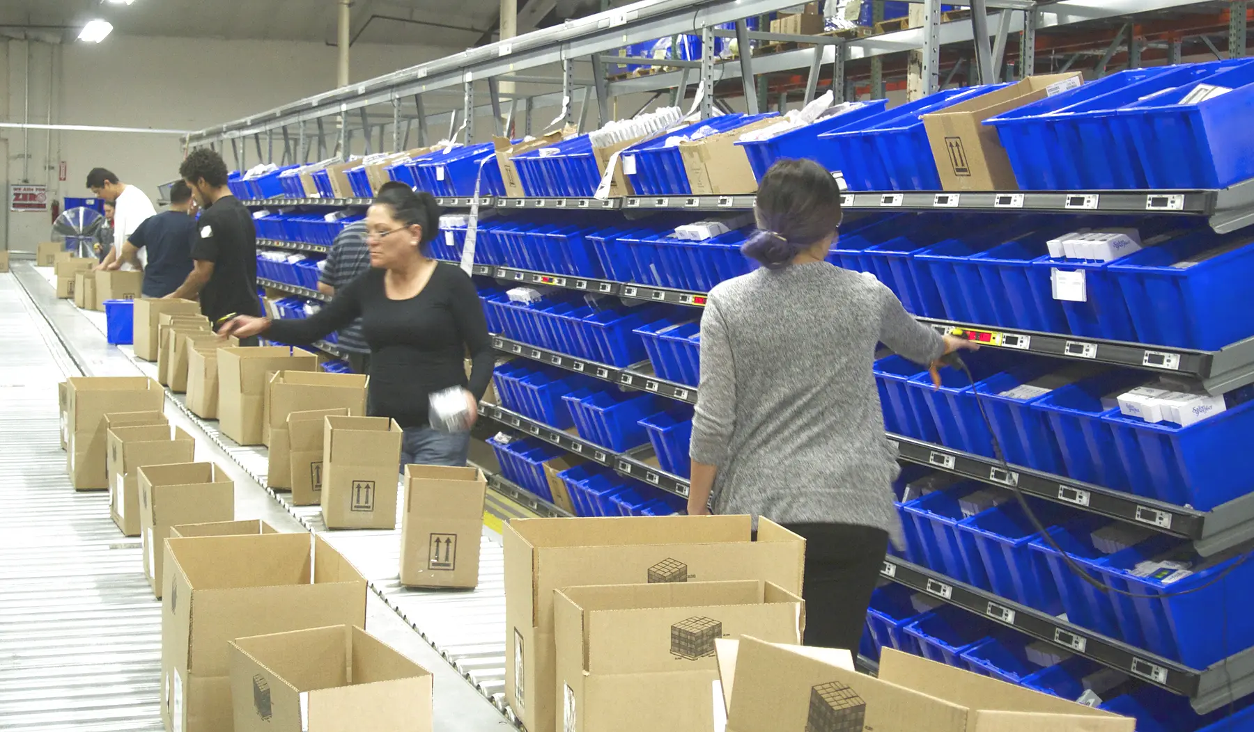 Operators picking from pick-to-light carton flow rack into shipping cartons moving along a takeaway conveyor in a fulfillment pick module
