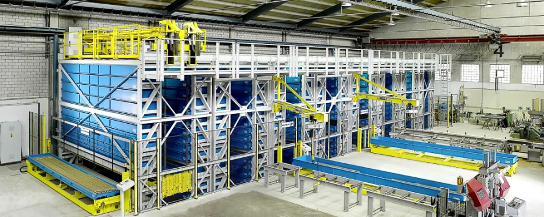 Long parts AS/RS cassette system with blue storage cassettes in a steel frame, gantry extractor, and outfeed transfer tables in a fabrication shop