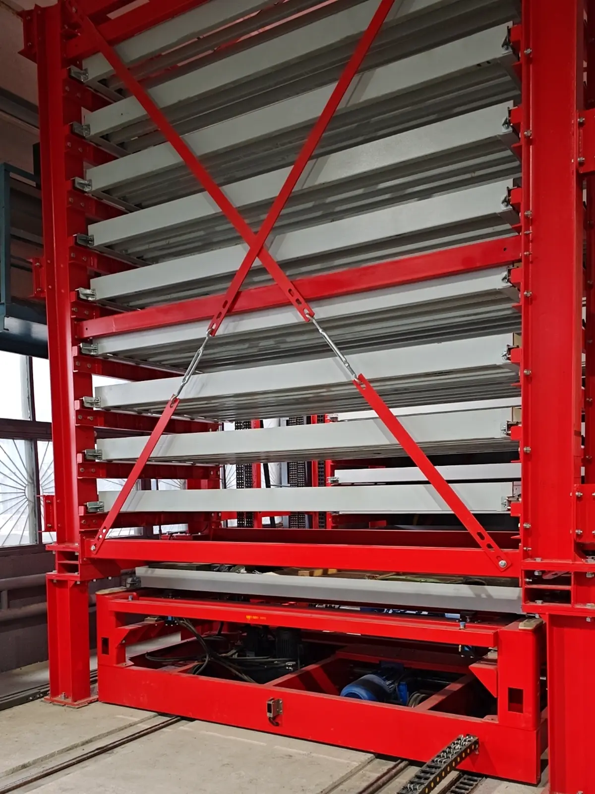 Long parts AS/RS tray tower with a red steel frame holding stacked storage trays above the extractor carriage and floor rails