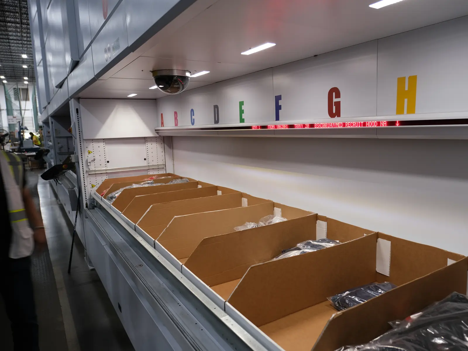 Vertical lift module batch pick station with an LED instruction bar, color-coded A through H put positions, and cardboard order bins staged at the tray access window